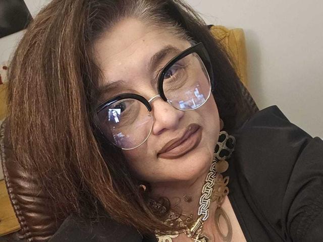 ClassybutNaughty with brown hair, glasses, and dark lipstick, wearing a black top and gold chain necklace, slightly smiling with a relaxed, winking expression.