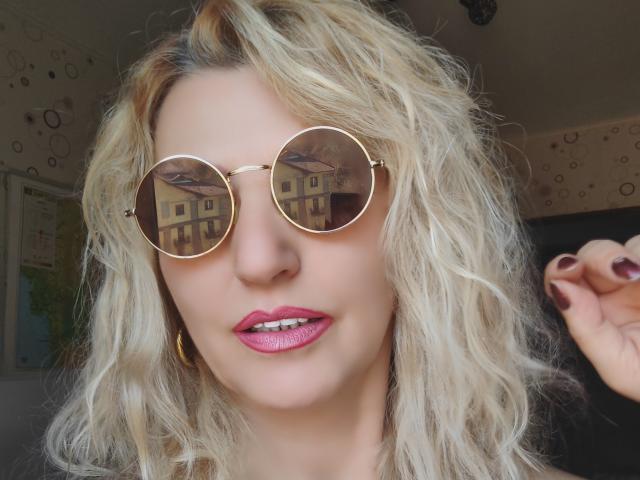 SexyCoco with wavy blonde hair, wearing round gold sunglasses, pink glittery lipstick, and gold hoop earrings, showing maroon nail polish, looking cute and stylish.
