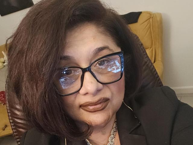 ClassybutNaughty with medium brown hair, black glasses, and brown lipstick, wearing a black jacket and silver chain, has a subtle smile and reflection in her glasses.