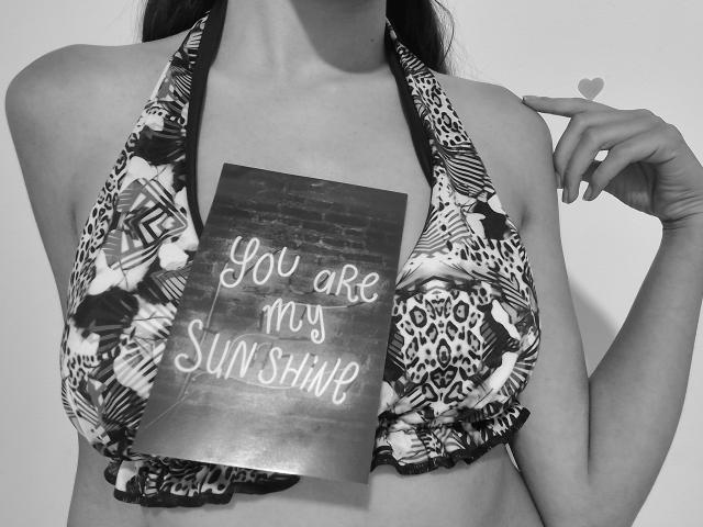 KiriamGomez with leopard print bikini, holding a sign over her tits, pointing with a heart-shaped finger, black-and-white photo, medium breast size, light skin, long dark hair.