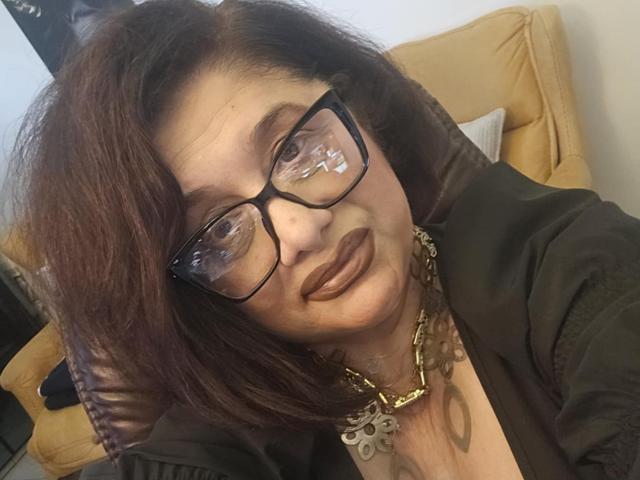 ClassybutNaughty with brown hair, glasses, and dark lipstick, wearing a black top and gold necklace, smiling slightly, head tilted to the side. Her tits are partially visible.