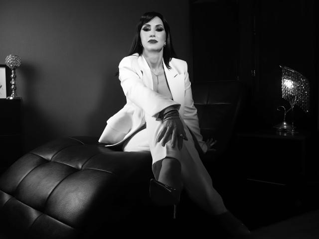 MissVonTease with dark hair, wearing a white suit, sitting on a black leather ottoman, her hand on her knee, looking confident, black heels, dark lipstick, black and white photo.