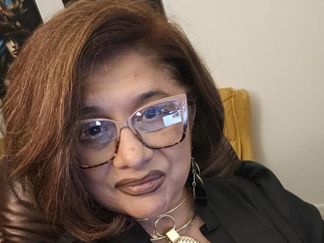 ClassybutNaughty has brown hair, wears clear glasses, and has brown lipstick. She's wearing a black shirt and a gold necklace with a heart-shaped pendant. Her ass is not visible.