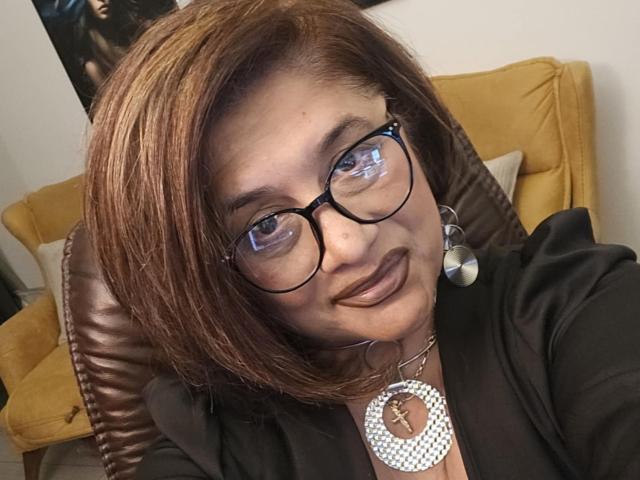 ClassybutNaughty with brown hair, glasses, and dark lipstick, wearing a black top and silver necklace with a large circular pendant, smiling at the camera.