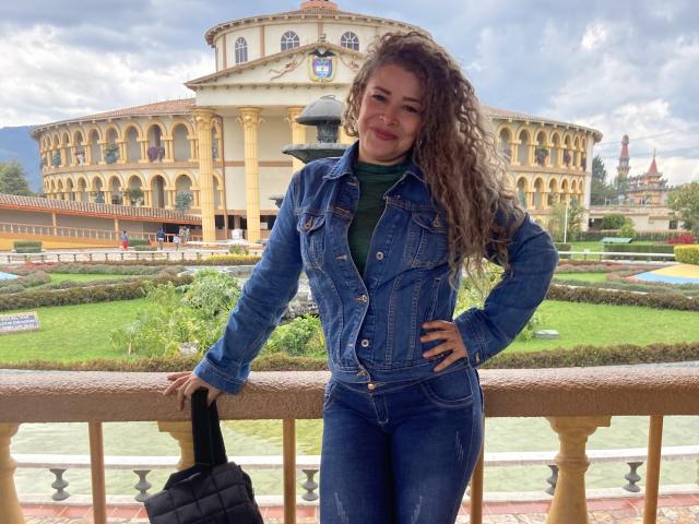 DalilaSuperHot with curly brown hair, wearing a blue denim jacket and green shirt, standing with hand on hip, holding black purse, smiling, in front of yellow building.