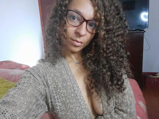 Angelonix with curly brown hair, wearing glasses and a grey knit cardigan, showing her tits in a deep V-neck, sitting on a pink couch.