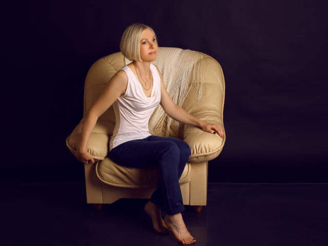 CityGlam with short blonde hair, white tank top, and blue pants sits barefoot on a beige leather chair, arms out, against a dark background.