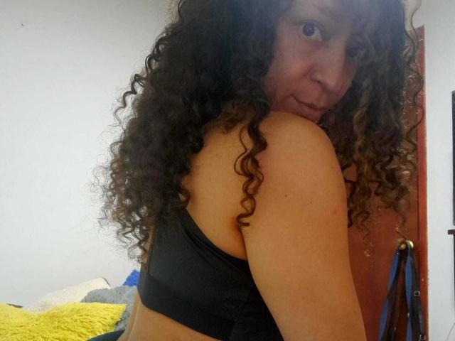 Angelonix with curly brown hair, black tank top, looking back, smiling, showing her shoulder and upper back, medium skin tone, bedroom setting.
