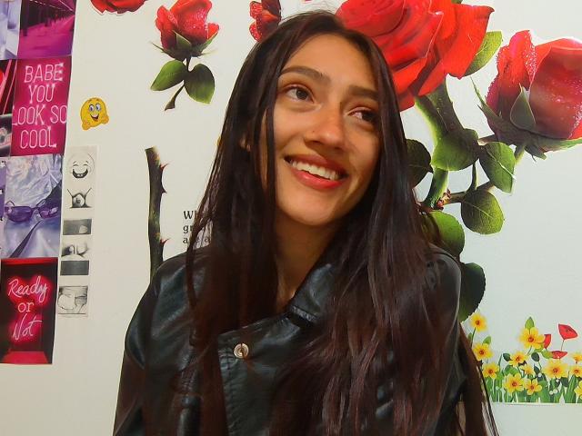 KiriamGomez, smiling with red lipstick, has long dark brown hair, wearing a black leather jacket, looking sideways with a playful expression, in a colorful, floral-themed photo.