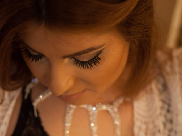 Sexydollhotx's face, brown hair, and closed eyes with long black eyelashes, looking down at sparkling white jewelry on her hands, wearing a lace top, soft warm lighting.
