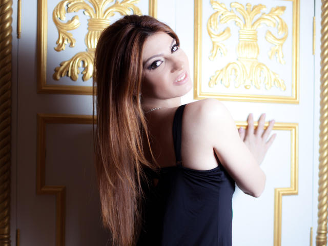 Sexydollhotx with long brown hair, black sleeveless top, looking back, smiling, holding door with gold ornate patterns, white and gold wall, showing bare shoulders and upper back.