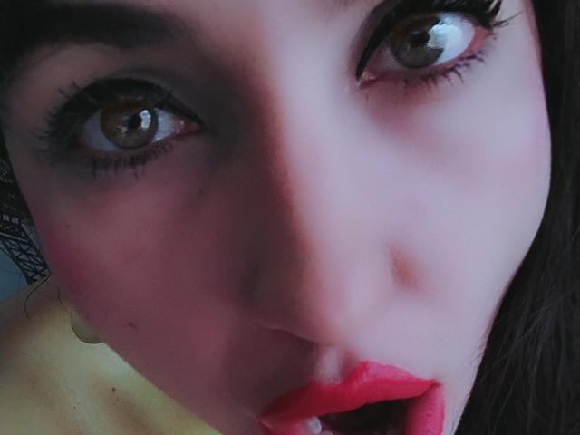 LuluFrancaise's face, showing brown eyes with thick black eyelashes, red lipstick, and a slight pink blush on pale skin. Her dark hair is partially visible on the right.