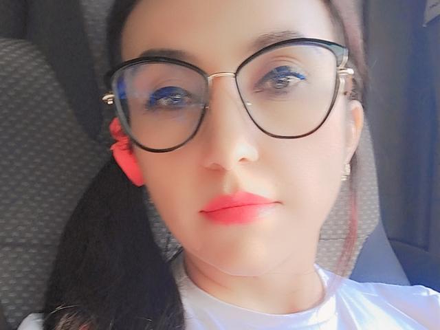 LuluFrancaise has pale skin, pink lipstick, blue eyeshadow, black glasses, and a pink hair tie. Her black ponytail is on the left, and she's wearing a white shirt.