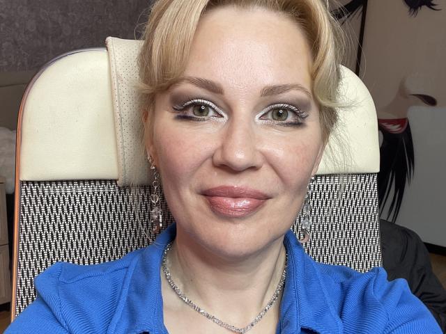 NatalyFoxy69 has blonde hair, green eyes, and is smiling with pink lips. She's wearing a blue shirt, silver necklace, and earrings, sitting in a chair with a white headrest.