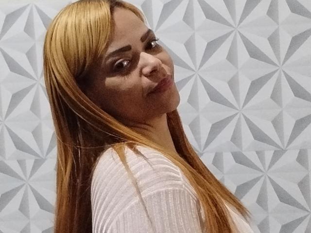 DeborahSweetX with long blonde hair, light brown skin, and a subtle smile, wearing a sheer pink top, looking over her shoulder. Her tits are slightly visible through the top.