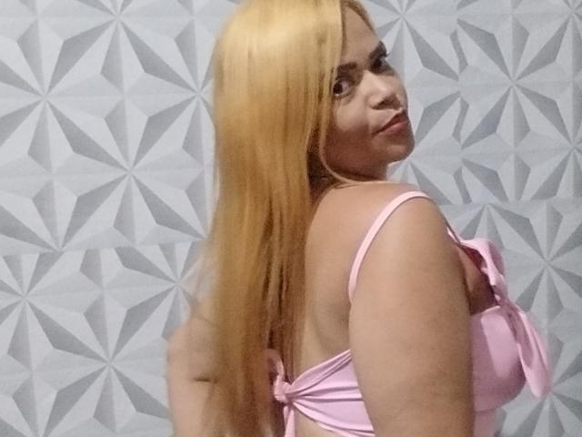 DeborahSweetX with long blonde hair, pink bra, and side glance, showing her back and tits, standing against a geometric patterned wall.