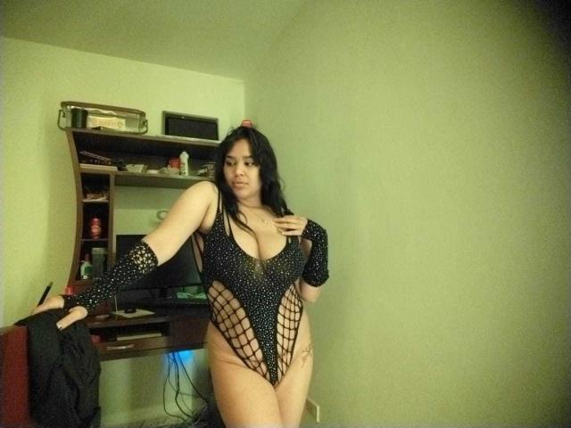 Shalyn with big tits, wearing a black, fishnet, high-cut, one-piece, revealing her pussy, standing in a room, leaning on a desk, long black hair, slight smile.