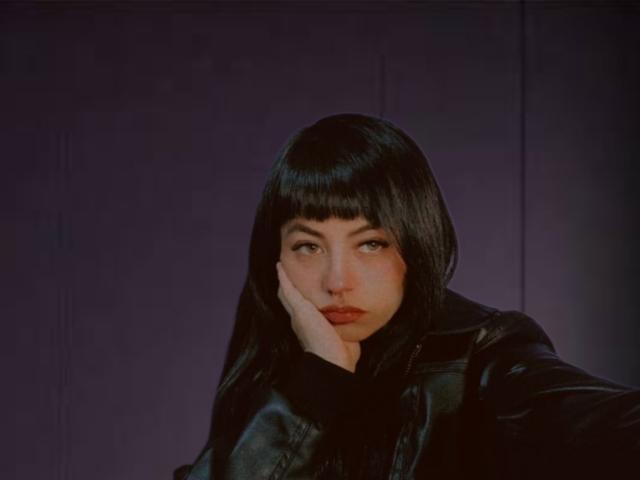 Cathulu with black bob, resting chin on hand, wearing black leather jacket, looking slightly to the side, has pale skin and dark eyes, against a dark purple background.