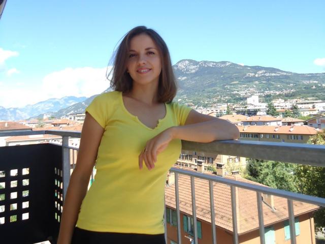 MissJoliSourireBg has brown hair, a yellow top, and black pants, leaning on a balcony with a mountain and town in the background, smiling with her hand on the railing.