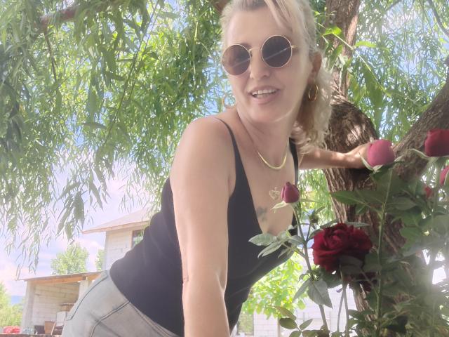 SexyCoco with blonde hair, wearing round sunglasses, black tank top, grey pants, gold necklace, leaning on tree, rose in hand, smiling, sunny outdoor setting, tree branches in background.