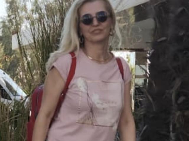 SexyCoco with blonde hair, wearing dark sunglasses, pink t-shirt with a graphic, red backpack, and a necklace, standing outside with a palm tree in the background.