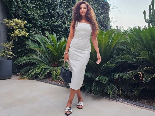 KayaOcean with curly brown hair, white strapless dress, black handbag, and white sandals, standing on a concrete path with green palm plants and a cactus.