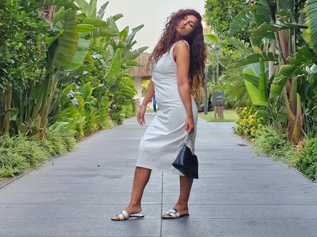 KayaOcean with curly brown hair, white sleeveless dress, black handbag, and white sandals, standing on a gray path, surrounded by lush green banana plants.
