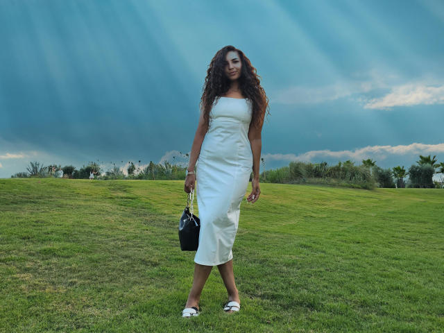 KayaOcean with long curly brown hair, wearing a white strapless dress, standing on green grass, holding a black purse, wearing white sandals, under a blue cloudy sky.