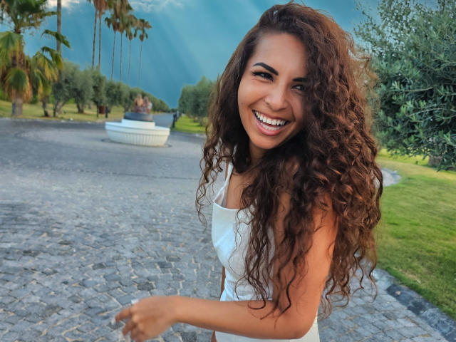 KayaOcean has long, curly brown hair, smiling with white teeth, wearing a white dress, standing on a cobblestone path, with green grass and trees in the background.