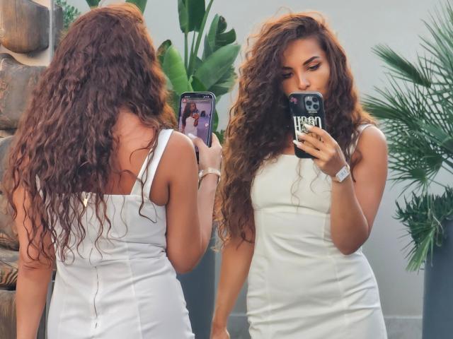 KayaOcean with long curly brown hair, wearing a tight white dress, taking a mirror selfie with her phone, showing her ass and small tits, in a sunlit room.