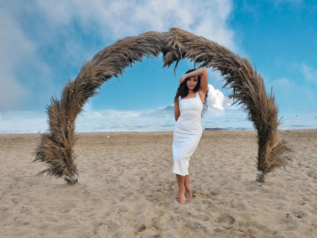 KayaOcean stands barefoot under a grassy arch, wearing a white, strapless, ankle-length dress, with one hand on her head, showcasing her ass and small tits. Beach, blue sky.