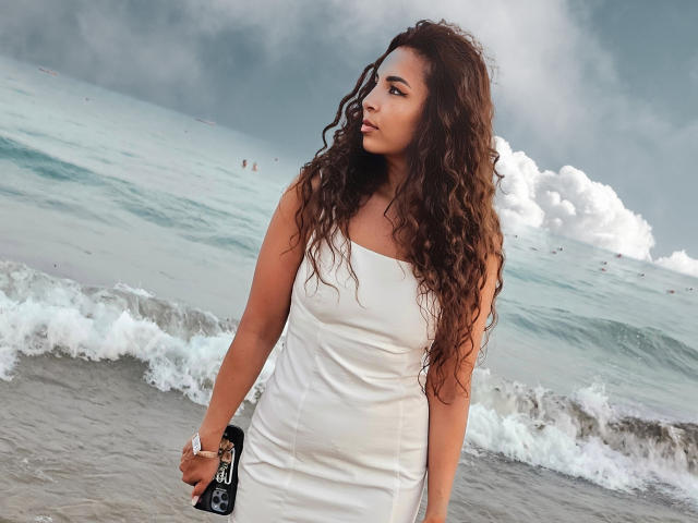 KayaOcean with curly brown hair in a white strapless dress, holding a black bag, standing on a beach with waves and cloudy sky. Her tits are medium-sized.