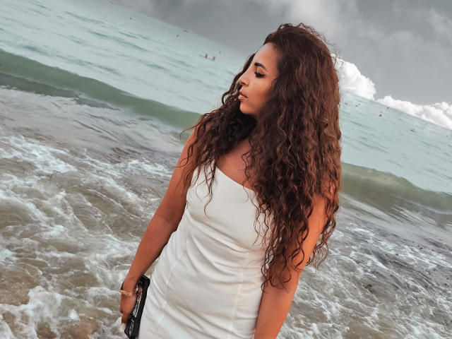 KayaOcean with long curly brown hair, wearing a white strapless dress, standing by the ocean, holding a black bag, looking to the side, under a cloudy sky.