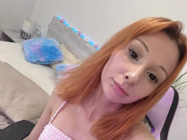 HannaCool has red hair, a nose ring, and a lip piercing. She's wearing a pink top, showing her small tits. She's sitting on a bed with a blue pillow.