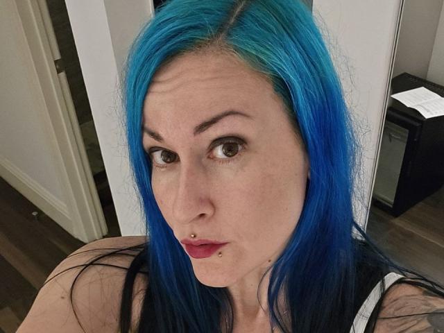 GennyRock with bright blue hair, nose piercing, and lower lip piercing, looking at the camera with raised eyebrows, wearing a black and white top.