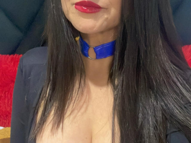 KynkyFetish with long black hair, red lipstick, and a blue choker, showing cleavage in a black top, focusing on her face and chest. Boobs visible, no other body parts.