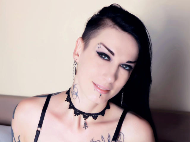 Evelinka-hot has black hair, tattoos, and piercings, wearing a lace choker and black straps, with blue eyes and a slight smile, looking at the camera.