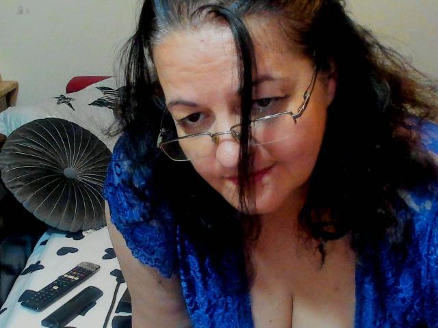 ReifeIri-hot with black hair, glasses, blue lace top, and visible cleavage, looking down with a slight smile. Remote control on bed.