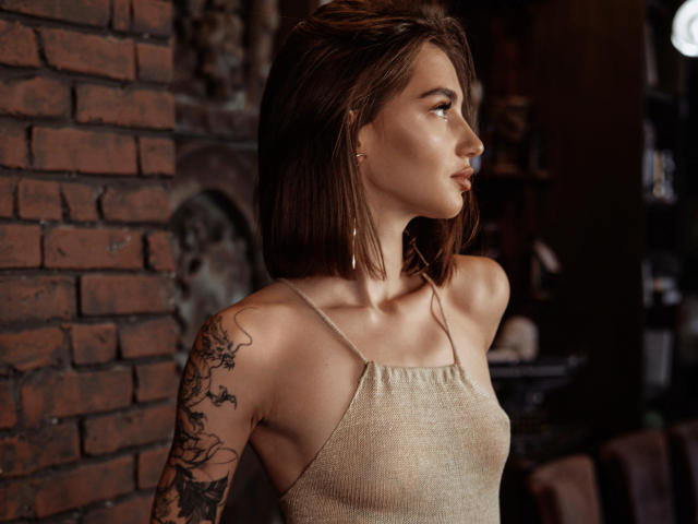 SophieKitty with shoulder-length brown hair, wearing a beige spaghetti-strap top, showing a tattoo on her right arm, standing near a brick wall, looking to the side.