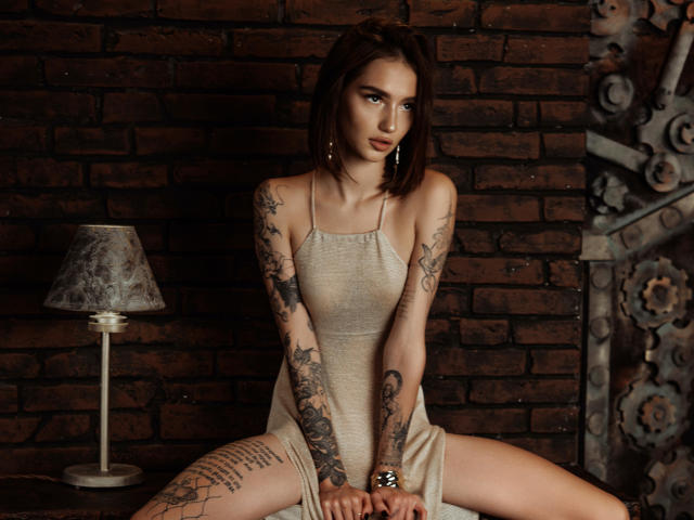 SophieKitty with tattoos, sitting on a surface, wearing a sheer, beige dress, legs spread, arms resting on thighs, looking at camera, brick wall in background, lamp to left.