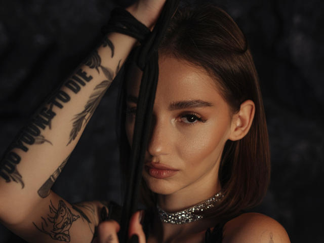 SophieKitty with brown hair, holding black fabric, has tattoos on her arm, a sparkly choker, and is looking seductively at the camera with her tits partially visible.