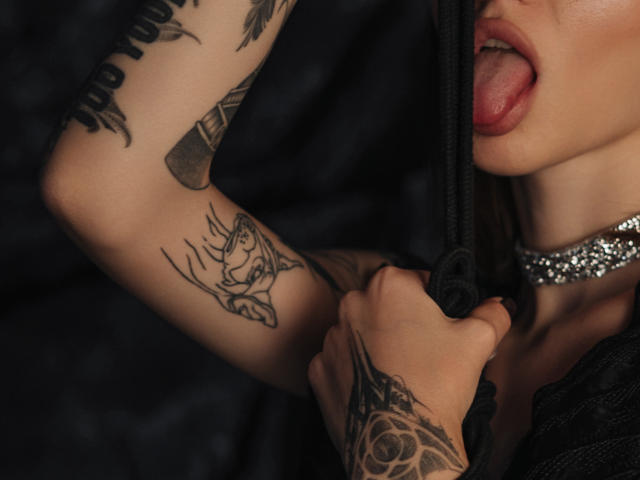 SophieKitty's tongue is out, biting black fabric, showing tattoos on her arm and hand. She wears a sparkly choker, with visible tattoos and her mouth slightly open.