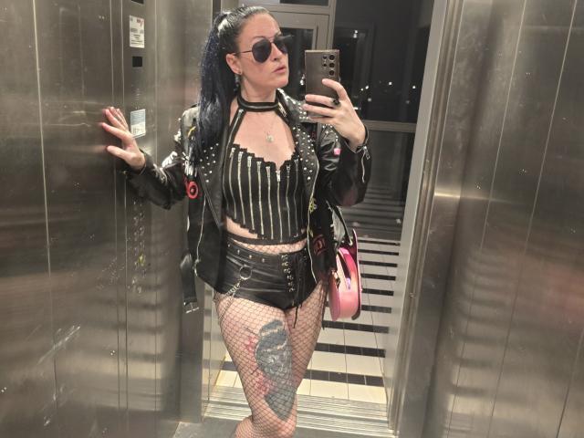 GennyRock with black pigtails, wearing a spiked leather jacket, striped top, black shorts, fishnet stockings, and a choker, taking a mirror selfie in an elevator. Her tattoo is visible.