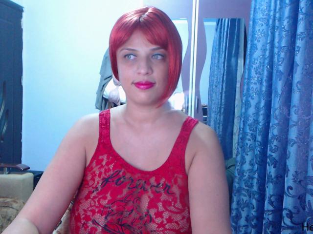 MyHairyCunt with red bob, wearing red lace tank top, showing tits, blue eyes, red lipstick, in a room with blue curtains, standing in front of a mirror.