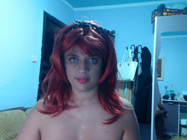 MyHairyCunt has red hair, blue eyes, and is topless with small tits. She's wearing a black hairband and has pink lipstick. The room has blue walls and a mirror.