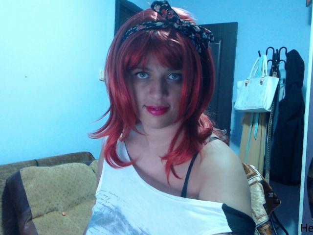 MyHairyCunt with red hair, black headband, white tank top, black straps, red lipstick, pale skin, blue eyes, smiling, in a room with a brown couch and white bag.