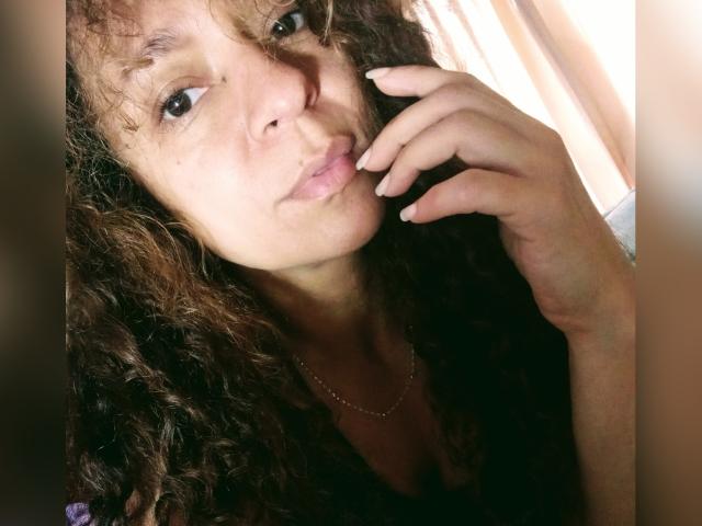 Angelonix with curly brown hair, light skin, pouty lips, and a subtle smile, touching her chin, wearing a thin necklace, and showing her small, cute tits.
