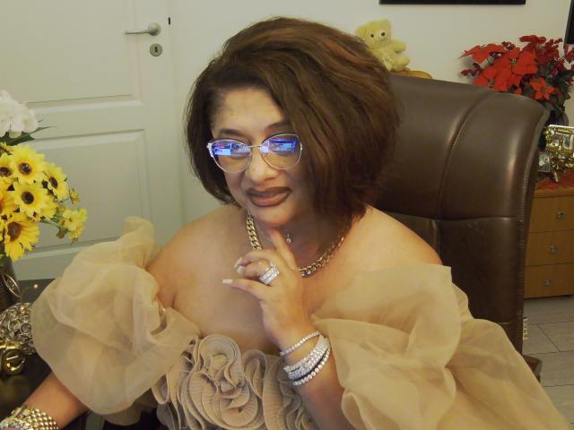 ClassybutNaughty with short brown hair, glasses, and a tan off-shoulder dress, showing her tits with a ringed finger, wearing jewelry, sitting in a brown chair.