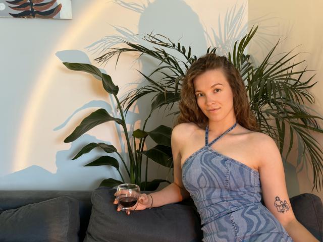 KenzieONEx with wavy brown hair, blue patterned halter dress, holding a glass of wine, tattoo on right arm, sitting on gray couch, green plants in background.
