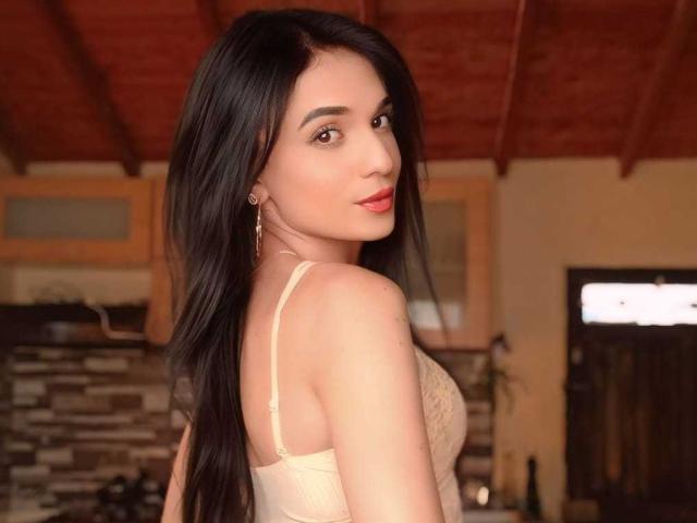 PamelaKander with long black hair, red lipstick, and a beige lace bra, showing her back and looking over her shoulder, has a cute butt and small tits.